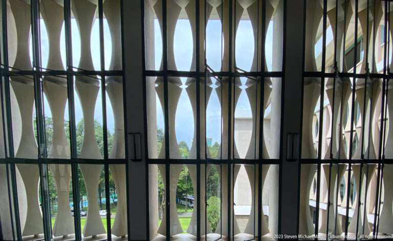 Close view through tall curved concrete screen modules toward trees and the palace grounds.