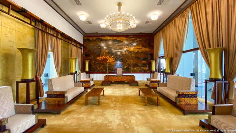 Centered Ambassadors’ Hall with cream sofas, gilded tables, tall vases, chandelier, and lacquer mural.