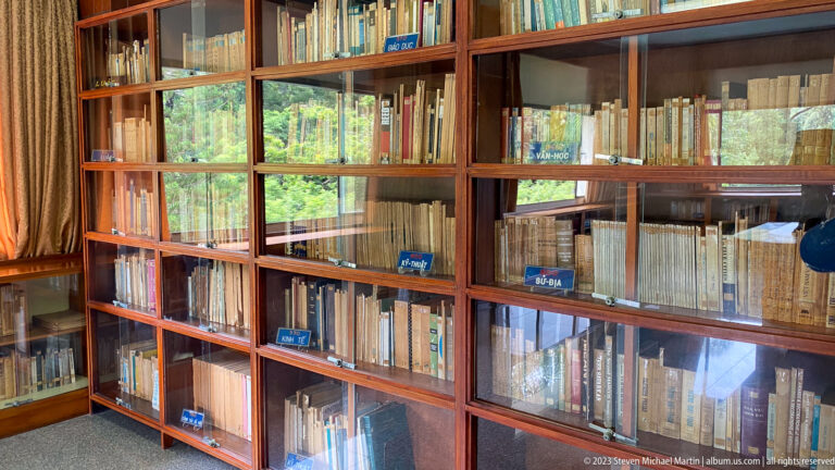 Glass-fronted wooden bookcases filled with Vietnamese-language books and small blue shelf labels.