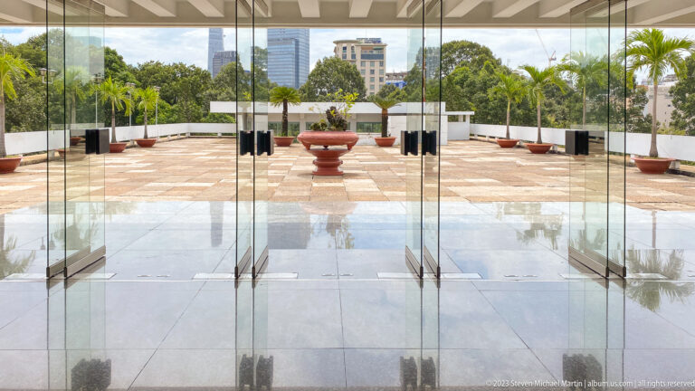 Open glass doors framing a tiled rooftop terrace with central planter, palms, and city buildings beyond.