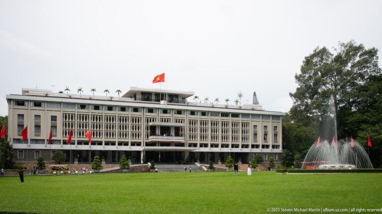 Wide view of Independence Palace across a broad green lawn, with the fountain to the right and flag above.