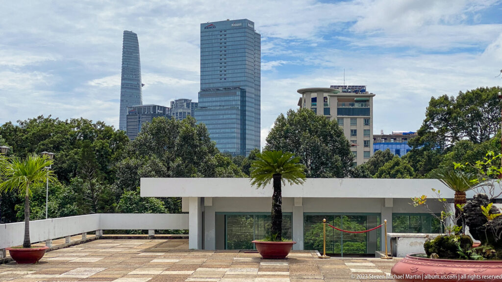 Palace rooftop terrace with white walls and potted palms facing modern city towers beyond trees.