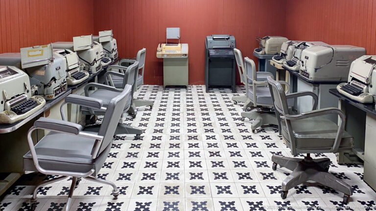 Narrow bunker room with rows of teleprinters, office chairs, patterned tile, and machines along both walls.
