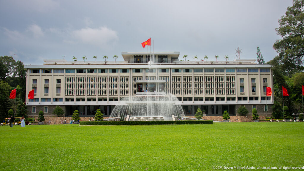 Centered frontal view of Independence Palace behind a fountain and broad lawn, with red flags at the sides.