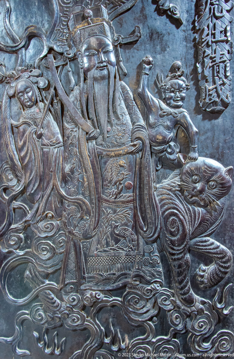 Silver-toned relief of a bearded robed figure riding a mythic animal amid attendants and Chinese text.