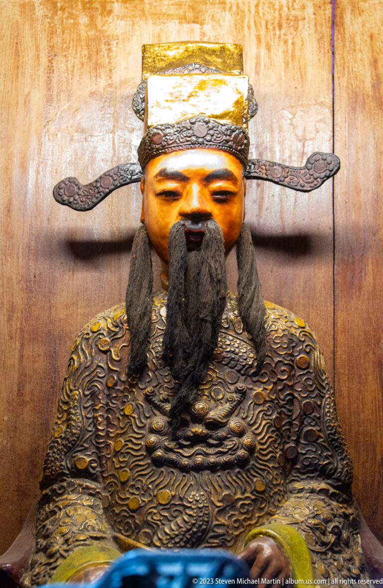 Seated bearded temple figure in patterned robes and a square headdress with long side projections.