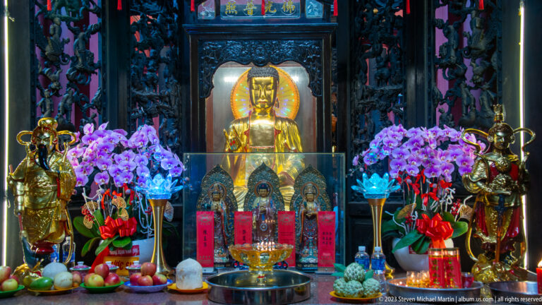Golden seated deity at a dark carved altar with orchids, fruit, incense, candles, and flanking figures.