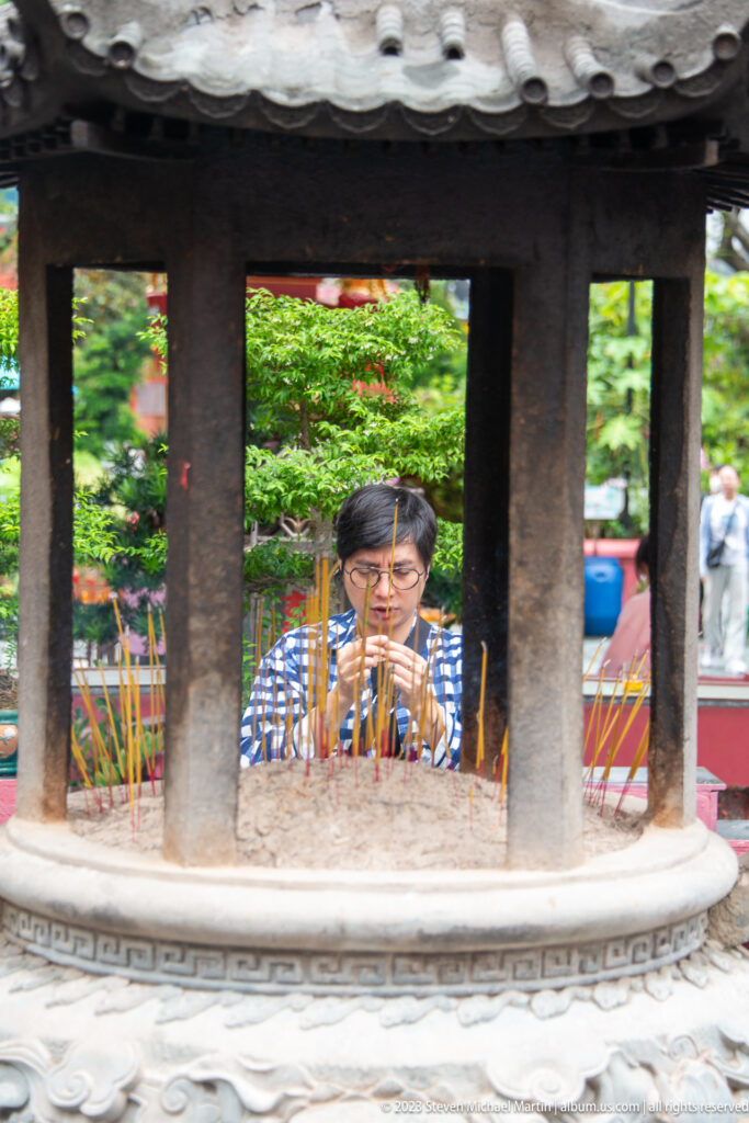 Worshipper seen through stone censer columns tending incense sticks in an ash-filled basin.