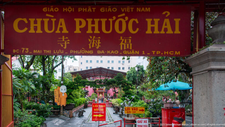 View beneath the Chùa Phước Hải sign into a leafy temple garden with statues and directional signs.