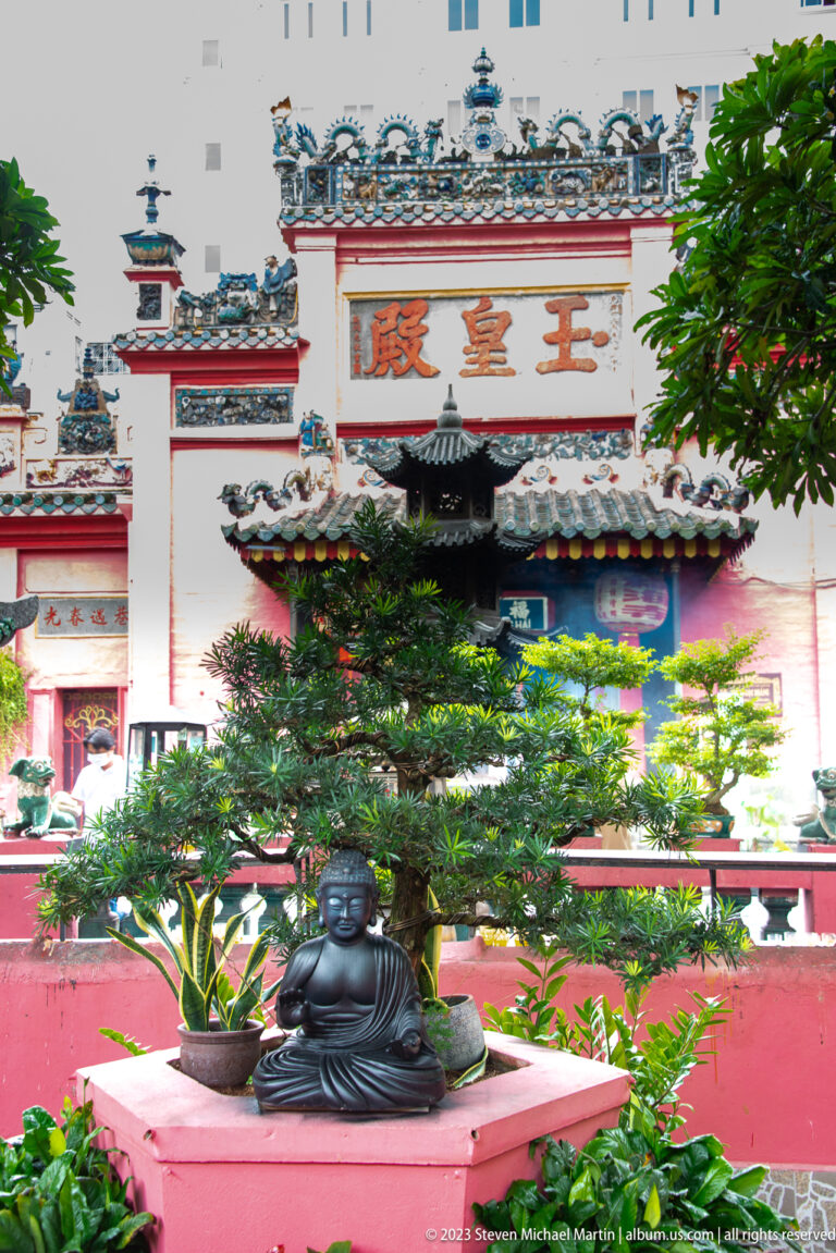 Pink temple gateway with Chinese inscription behind a seated dark Buddha figure and green courtyard plants.