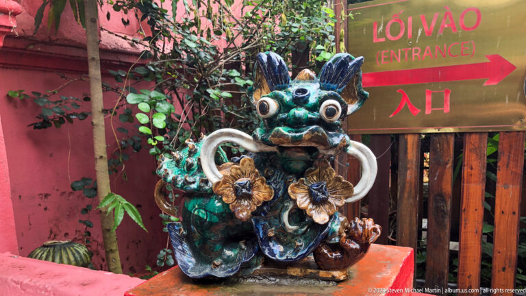 Multicolored glazed guardian creature beside a gold entrance sign in Vietnamese, English, and Chinese.