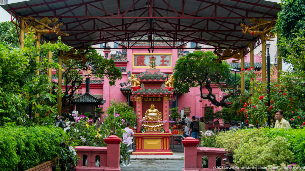 Covered temple courtyard with a small red shrine, golden figure, clipped trees, and pink walls.
