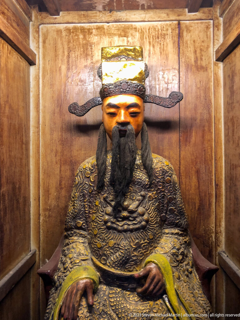 Seated bearded temple figure in patterned robes and projecting ceremonial headgear inside a wooden niche.