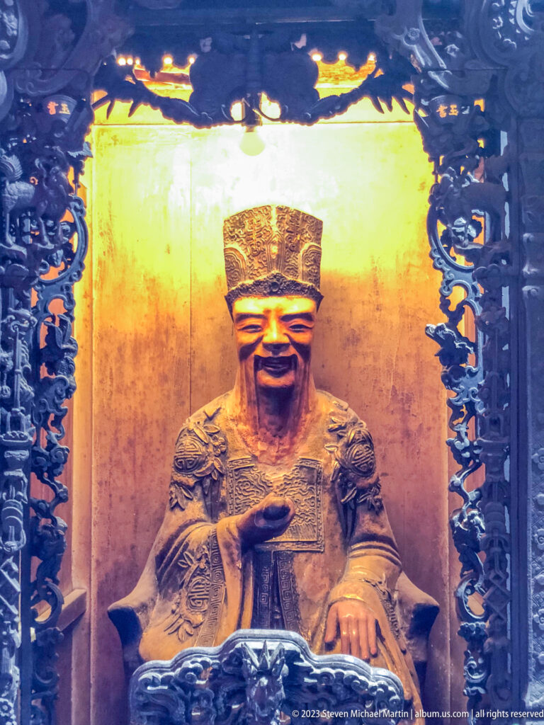 Seated bearded temple deity in a tall headdress and patterned robes inside a dark carved shrine.