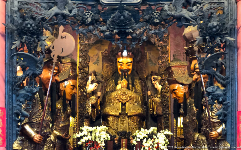 Central enthroned deity with armed guardian figures, carved dragons, and white flower offerings at a temple altar.