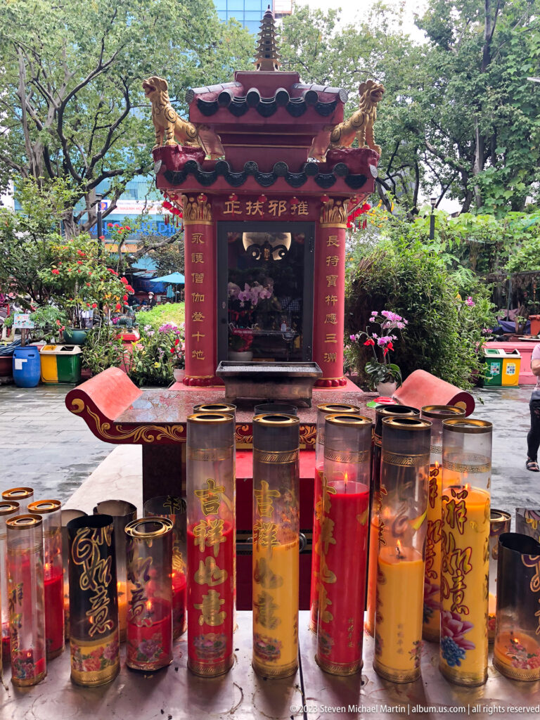 Rows of decorated cylindrical incense containers standing before a small red shrine in a leafy courtyard.