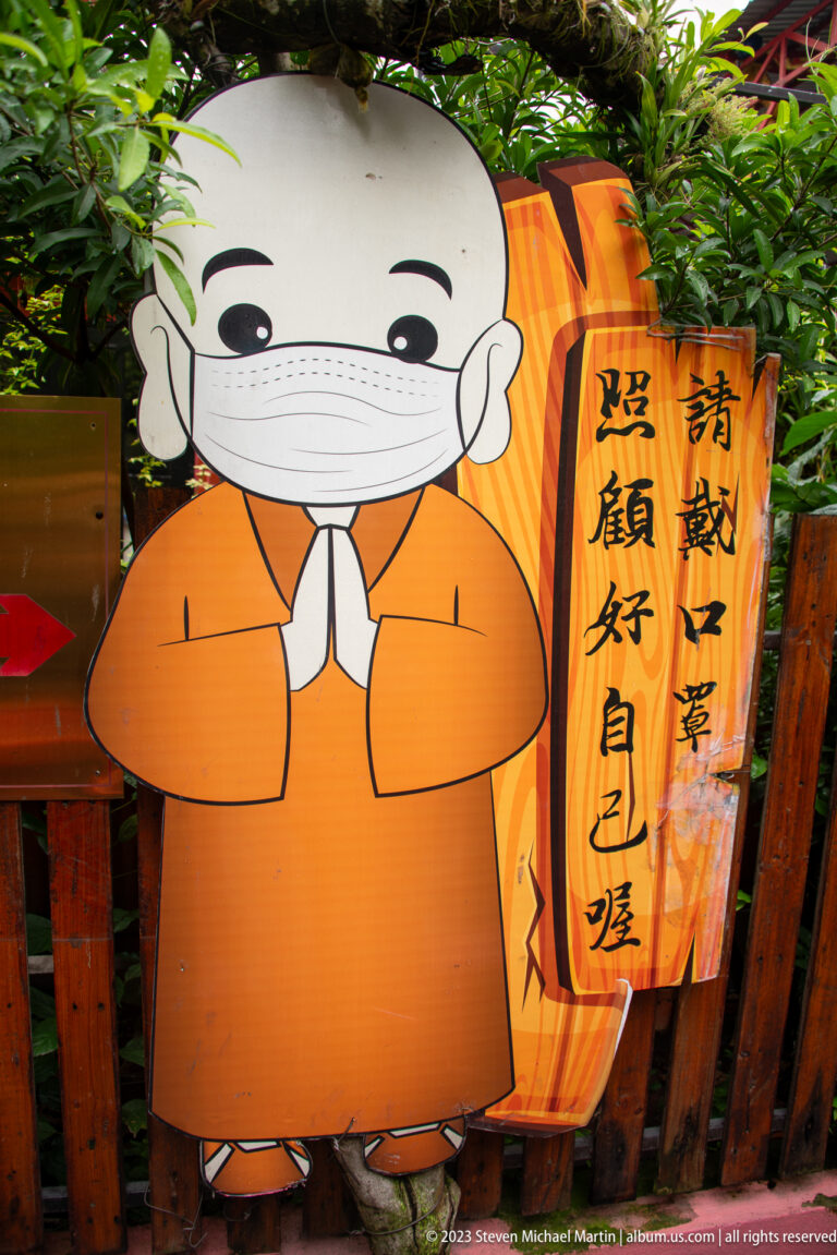Cartoon child monk in orange robes and a face mask beside a vertical Chinese calligraphy panel.