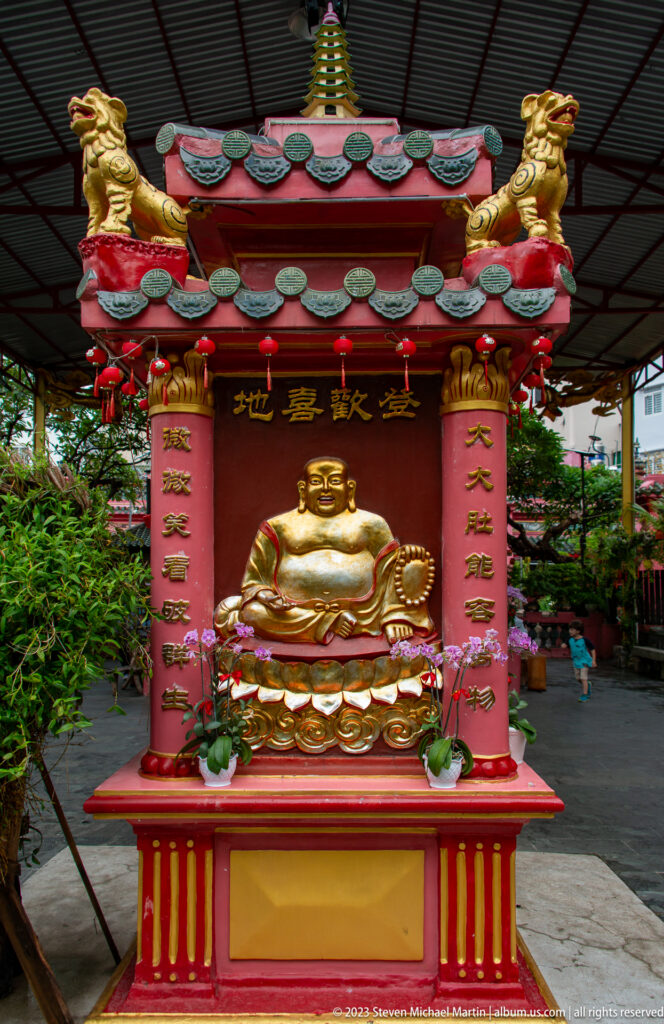 Golden laughing Budai figure seated in a red and gold pavilion beneath a tiled roof.