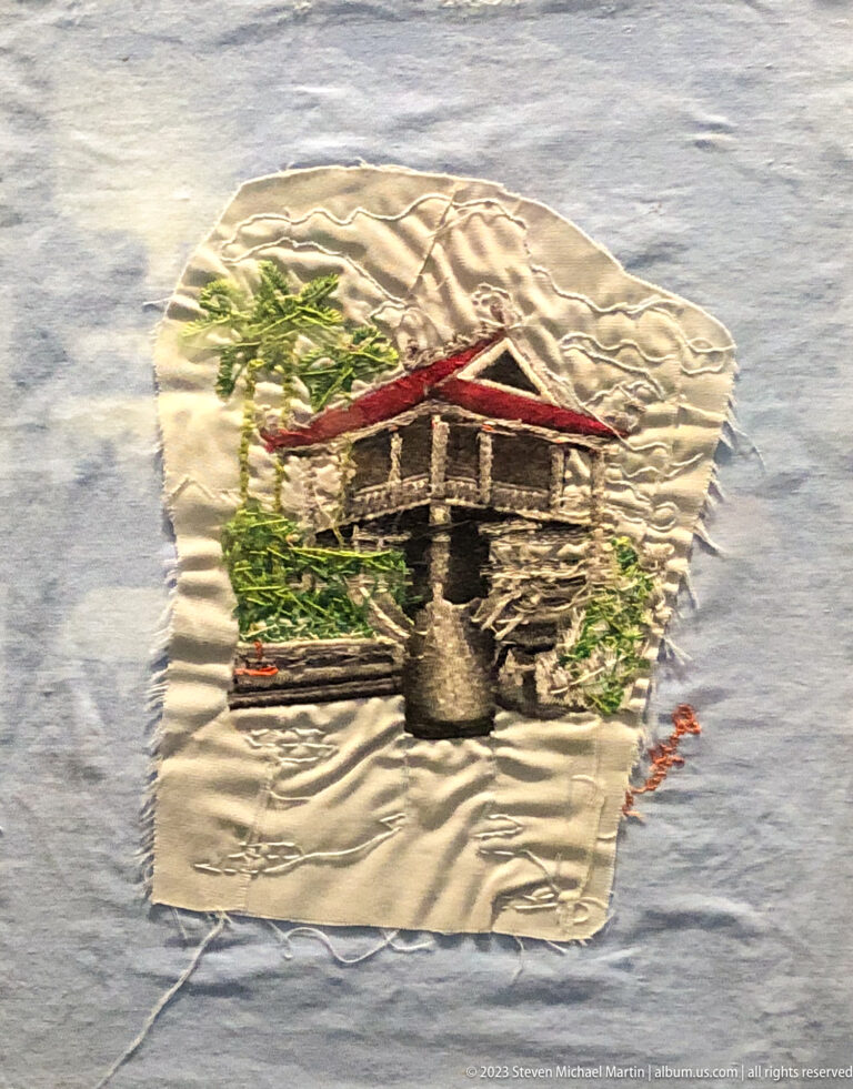 Embroidered stone building with red roof and greenery on pale fabric.