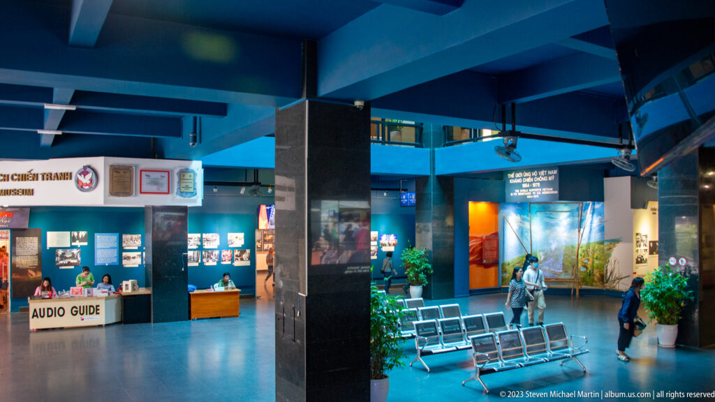 Blue museum lobby with seating, audio-guide desk, and exhibition entrances.