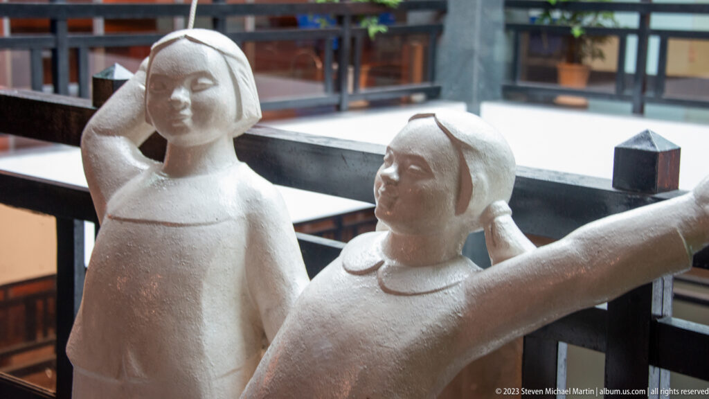 Two pale sculpted women beside a museum railing, one with arms extended.