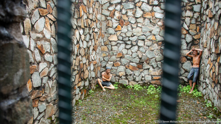 Two prisoner mannequins inside a narrow stone-walled enclosure viewed through bars.