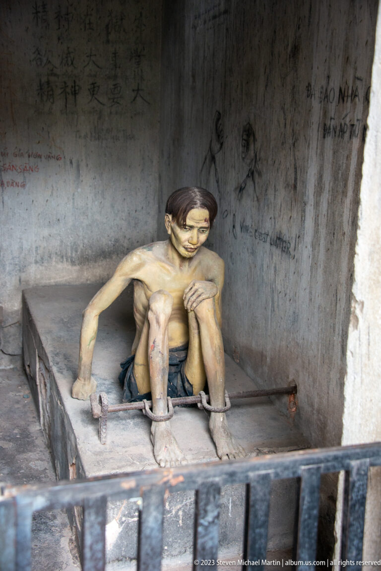 Gaunt prisoner mannequin seated on a concrete platform inside a cell.