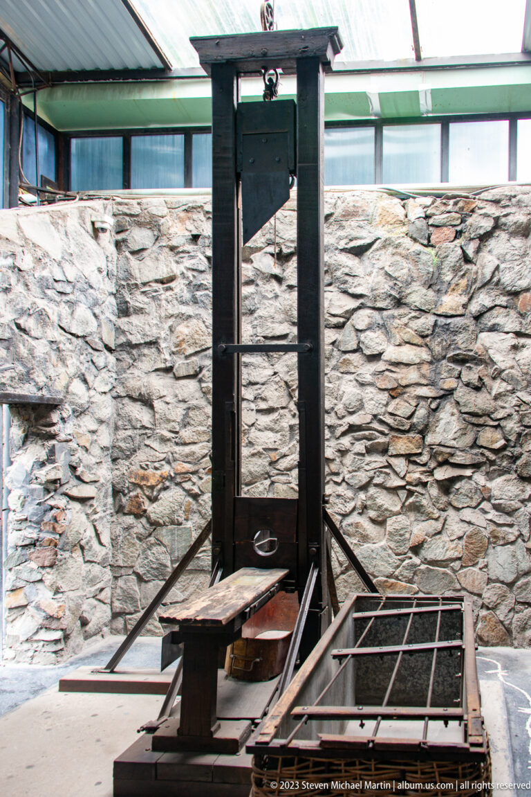 Guillotine with tall frame and wooden platform in a stone-walled courtyard.