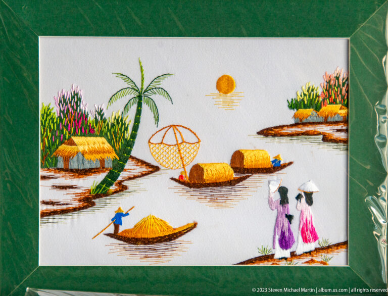Framed embroidery of a Vietnamese river village with boats and palm trees.