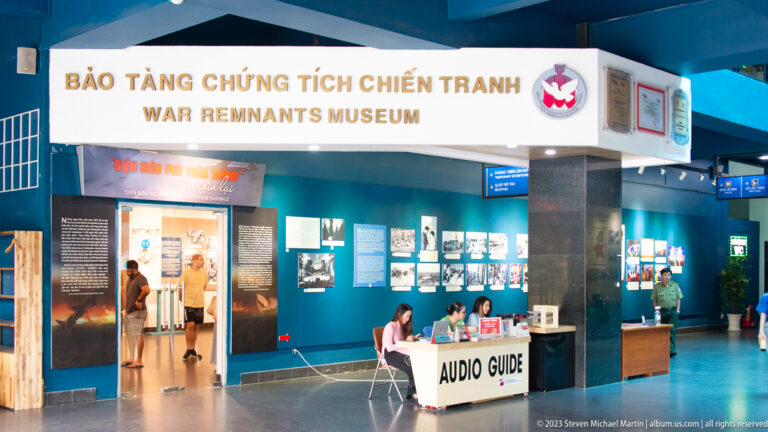 Museum lobby with bilingual sign, audio-guide desk, visitors, and exhibition panels.