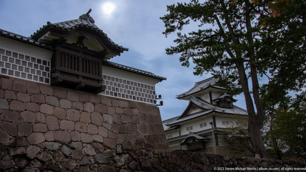 Kanazawa Castle turret and white wall rising above a massive stone rampart under trees.