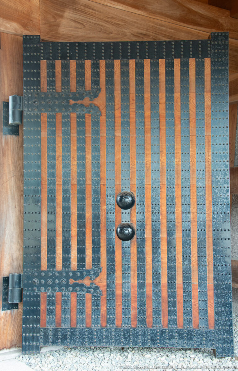 Closed wooden castle gate covered with iron straps, studs, hinges, and central metal fittings.