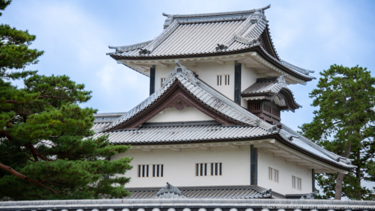 White Kanazawa Castle turret with layered gray tile roofs framed by pine trees.