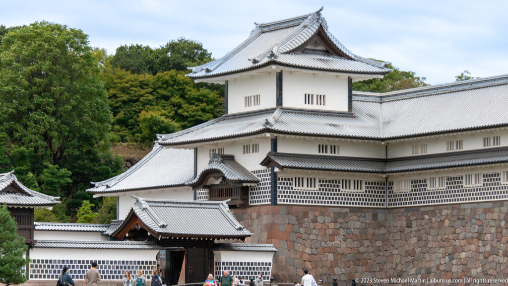 Large white Kanazawa Castle complex with tiled roofs, stone base, gateway, and visitors below.