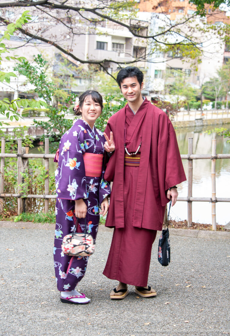 Couple in purple and burgundy kimono posing beside a landscaped pond and bamboo fence.