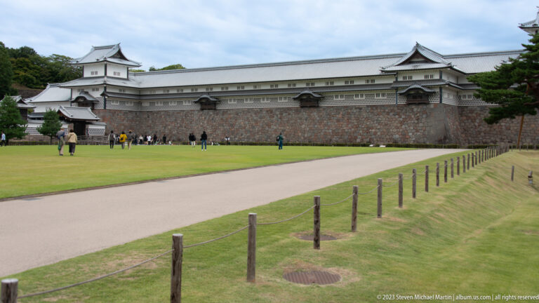 Long white Kanazawa Castle building on a high stone base beside a broad lawn and path.