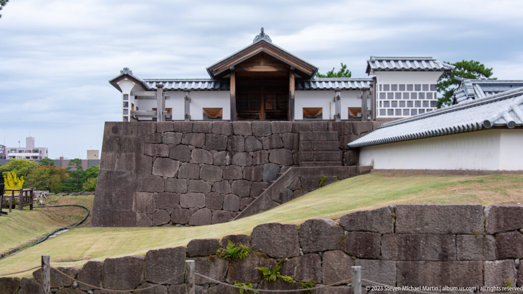 White castle gate above massive stepped stone walls and sloping grass at Kanazawa Castle.