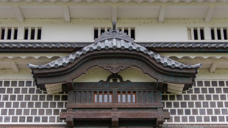 Ornate projecting timber lattice window beneath a curved gray tile canopy on a white wall.