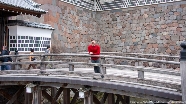 Man in a red shirt standing on a curved wooden bridge beside high Kanazawa Castle stone walls.