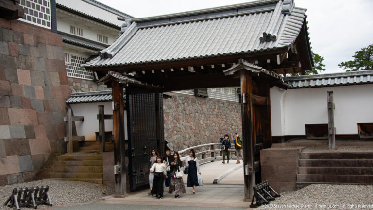Open timber gate between white plaster and stone walls, with visitors passing through.