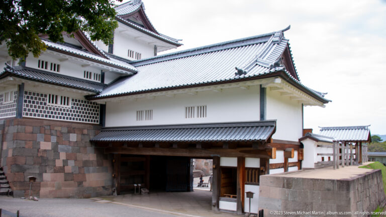 Covered gateway beneath white Kanazawa Castle buildings with layered gray tile roofs and stone base.