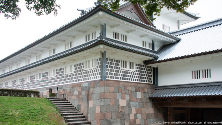 White castle corner with layered gray roofs and dark lattice panels above a colorful stone base.
