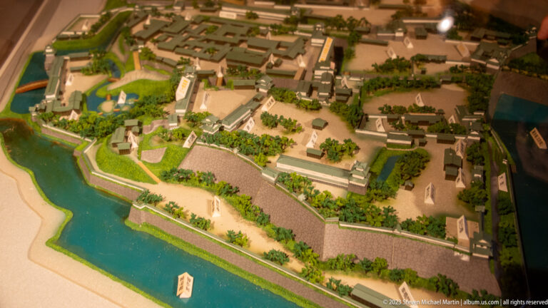 Overhead view of a scale model showing Kanazawa Castle buildings, moats, stone walls, paths, and landscaped grounds.