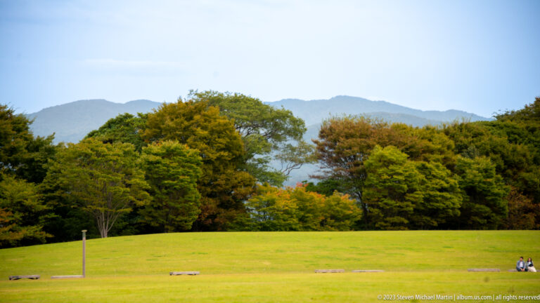 Wide green lawn bordered by mature trees, with hazy mountains beyond Kanazawa Castle Park.