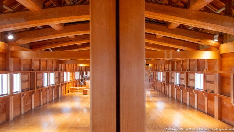 Large reconstructed wooden interior with a central post, exposed beams, braces, polished floorboards, and rows of windows.