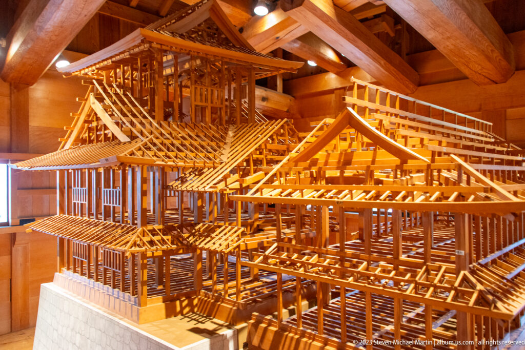 Oblique view of an extensive wooden castle model with intersecting roof frames, posts, corridors, and stone bases.