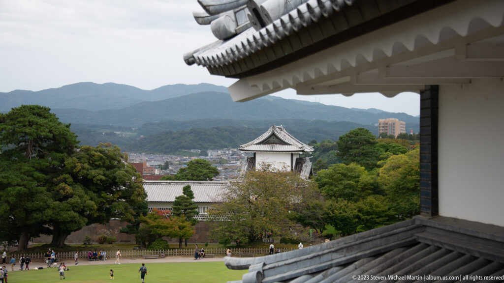 White castle eaves frame a view of a turret, lawn, trees, city buildings, and wooded hills beyond Kanazawa Castle.