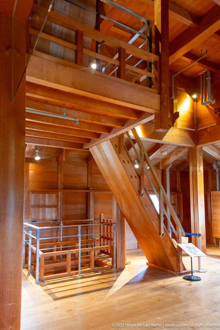 Wooden stair rising through an interior of heavy posts, beams, floor framing, railings, and interpretive displays.
