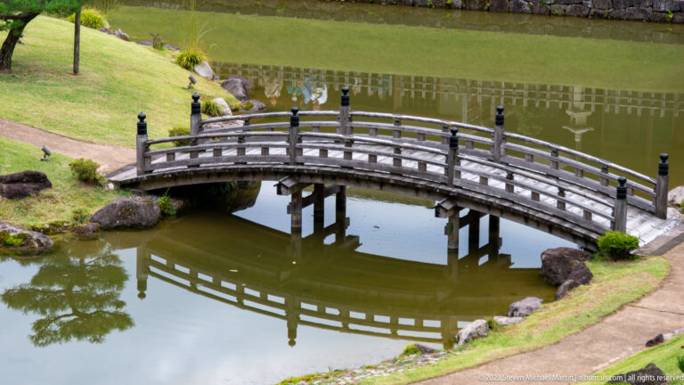 Arched wooden bridge crossing a reflective pond amid rocks, clipped shrubs, and grassy banks in a formal garden.