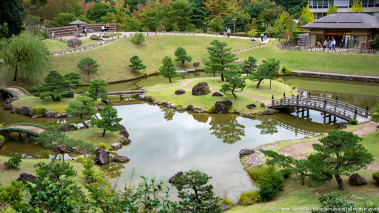 Elevated view of a Japanese garden with pond, two bridges, clipped pines, rocks, paths, and a pavilion.